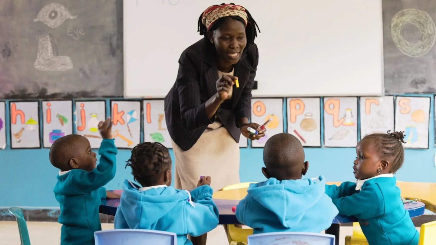 A teacher leading a lesson as children around the table raise their hands.