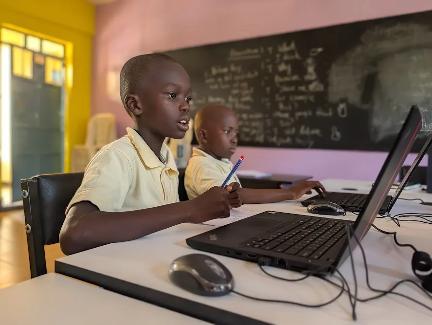Two DUMA pupils at laptops, with a pencil and written work beside the computers.