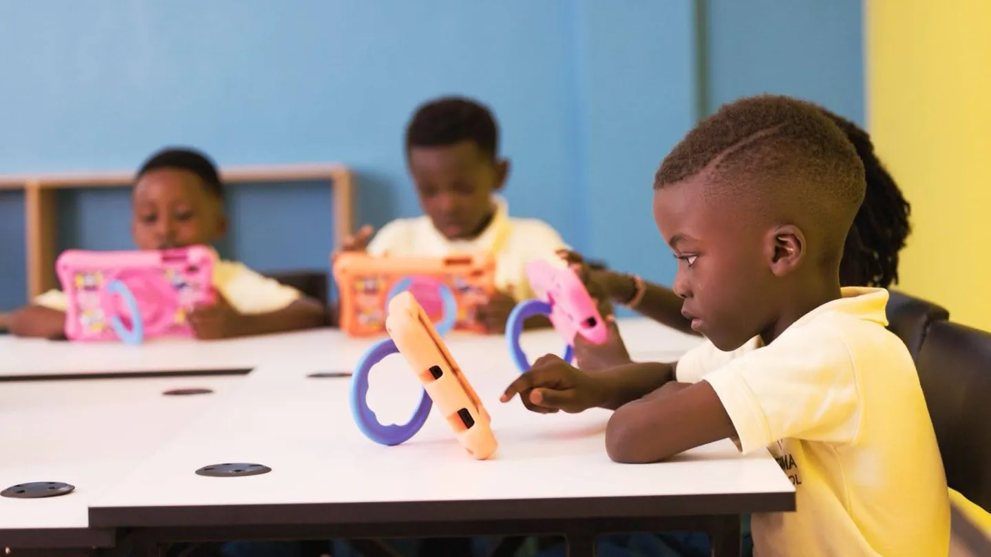 Pupils using colourful tablets at a white classroom table, with one child in focus in the foreground.